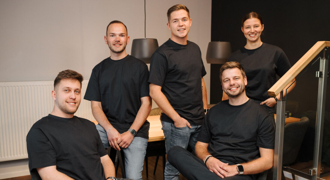 Fünf junge Erwachsene in schwarzer Kleidung posieren gemeinsam in einem modernen Büroambiente und lächeln in die Kamera.