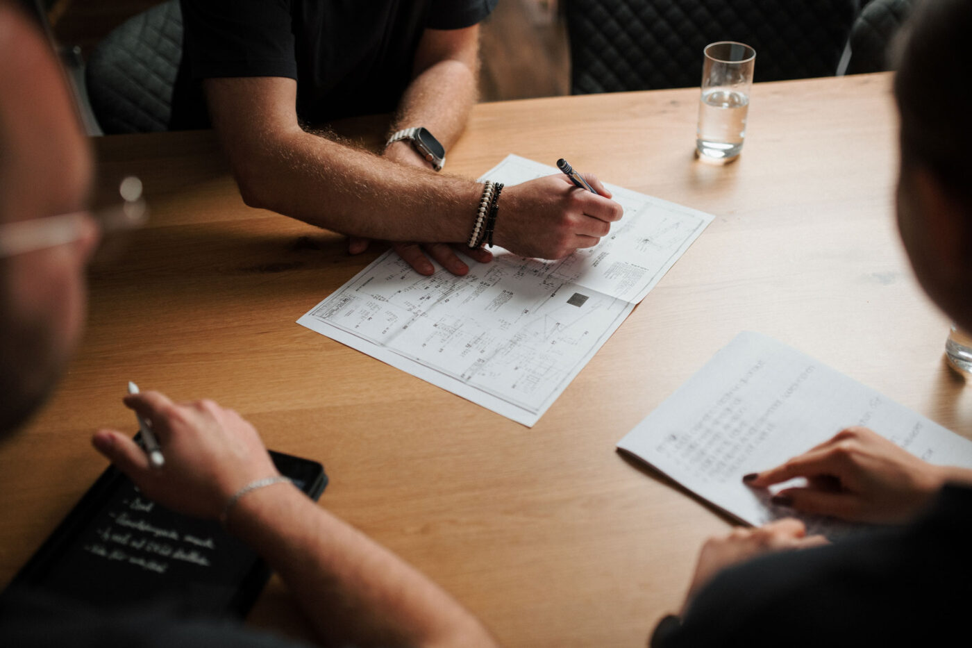 Wir wir arbeiten : Hände mehrerer Personen bei einer Besprechung, die technische Zeichnungen und Projektunterlagen auf einem Holztisch prüfen.