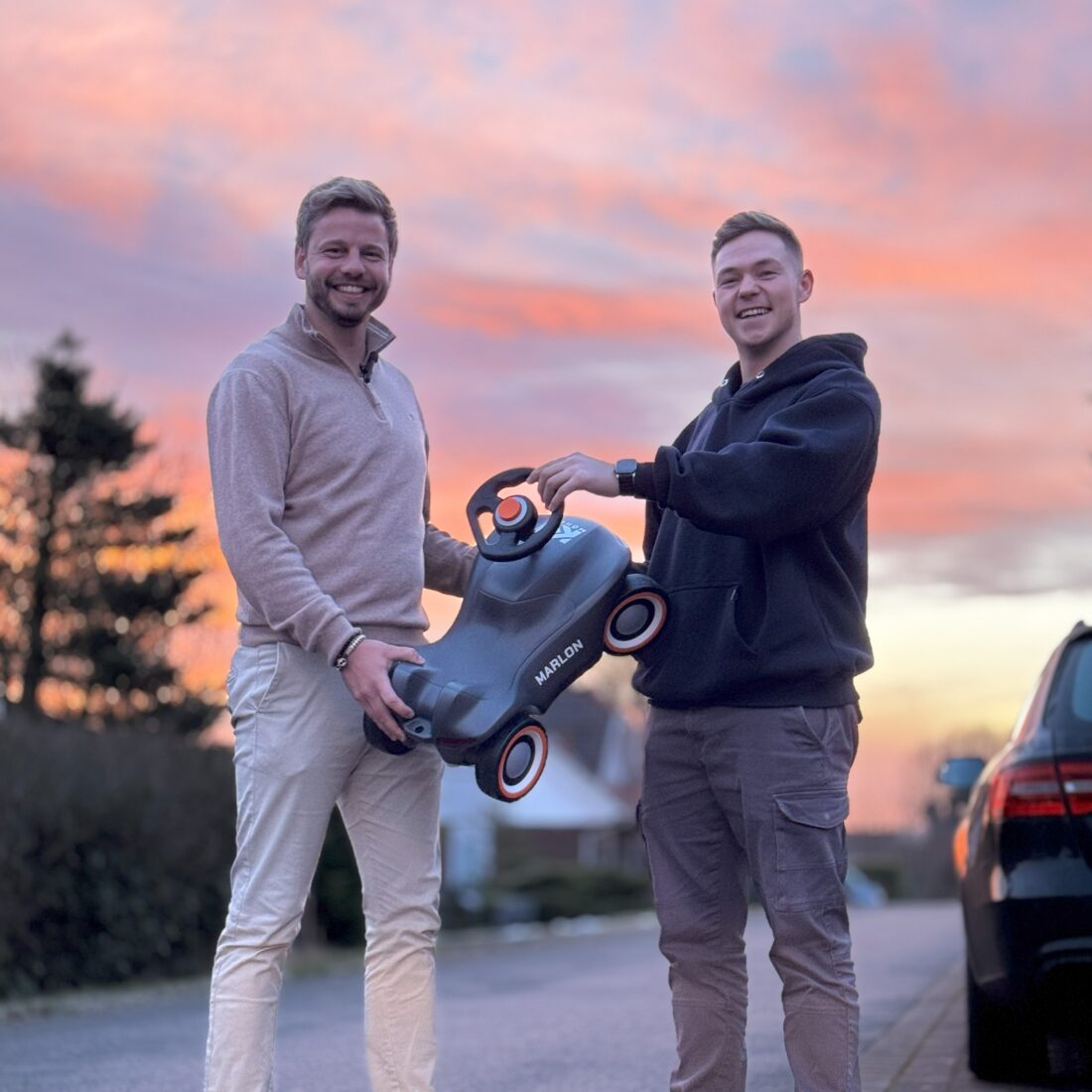 Zwei lächelnde Männer halten gemeinsam ein Kinderfahrzeug bei Sonnenuntergang auf einer Wohnstraße.