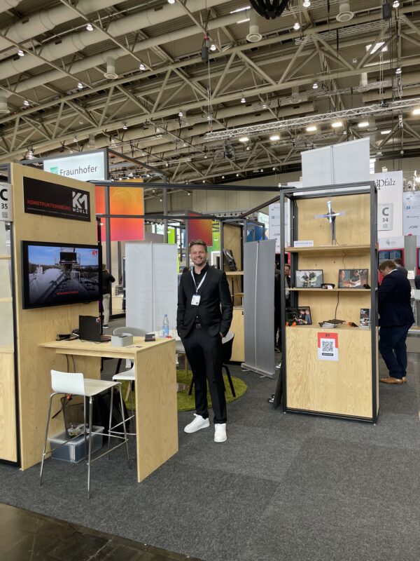 Mann im schwarzen Anzug am Stand von Konstruktionsbüro MÖHLE auf der Hannover Messe 2025, umgeben von Displays, Prospekten und einem Bildschirm mit Präsentation.