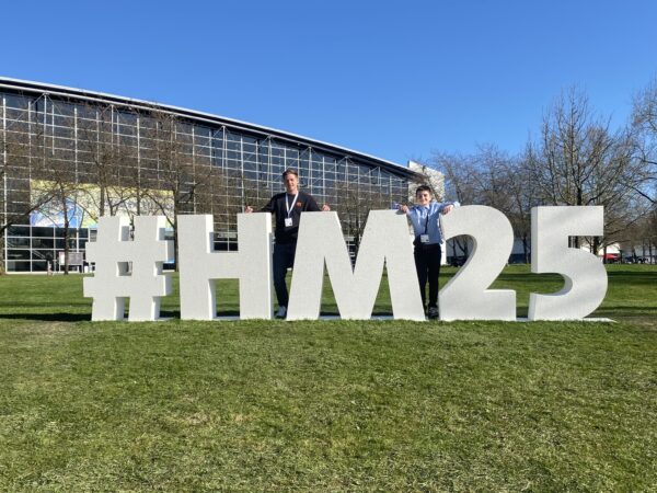 Zwei Personen posieren im Sonnenschein vor einer großen weißen #HM25-Installation auf dem Außengelände der Hannover Messe.