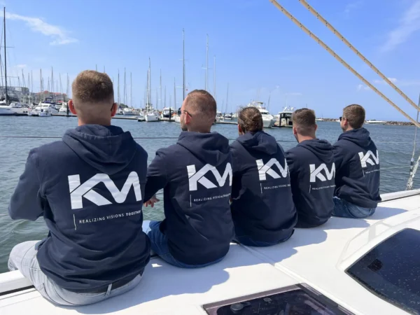 Fünf Männer in einheitlichen KM-Hoodies sitzen auf einem Boot und blicken auf einen Yachthafen mit vielen Segelbooten.