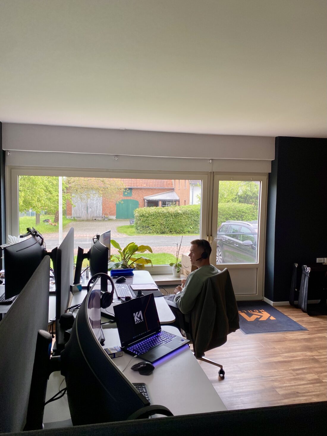 Mitarbeiter mit Headset sitzt in einem modernen Büro mit mehreren Arbeitsplätzen und Curved-Monitoren, vor einem großen Fenster mit Blick auf eine ruhige Wohnstraße.