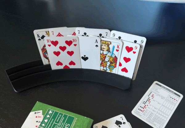 Ein Skat-Spiel ist im Gange: Auf einem dunklen Holztisch stehen Spielkarten in einem schwarzen Kartenhalter. Im Vordergrund liegen eine grüne Skat-Kartenverpackung, einige abgelegte Karten sowie ein Skatblock mit Punkten. Im Hintergrund befinden sich weitere Alltagsgegenstände.